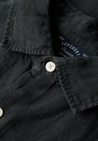 Camicia nera in lino con colletto a punta, dettaglio in bianco con bottone e etichetta blu con la scritta "Superdry Premium". Texture liscia.