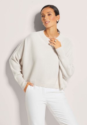 Helles beiges Oversize-Pullover mit Rundhalsausschnitt, geripptem Saum und überschnittenen Schultern, kombiniert mit weißen Hosen. Weicher, strukturierter Stoff.