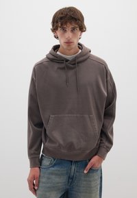 Felpa marrone in cotone con tasca frontale, cappuccio con cordino e polsini a costine. Abbinata a jeans in denim chiaro.