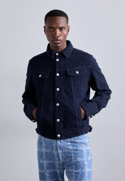 Veste texturée bleu marine avec des boutons argentés, dotée de deux poches poitrine et d'un col, associée à un jean bleu clair arborant un texte.