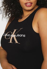 Svart bomullstanktop med en stor beige "CK"-logotyp och texten "Calvin Klein Jeans". Designen inkluderar en ärmlös skärning och ribbad halsringning.