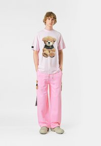 Camisa rosa de gran tamaño con un gráfico de oso de peluche, combinada con pantalones rosa a juego que presentan acentos de oso de peluche. El modelo lleva calzado de color claro.
