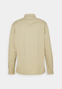 Chemise beige à manches longues vue de dos avec un col, des poignets boutonnés et une petite boucle en tissu sur le haut du dos.