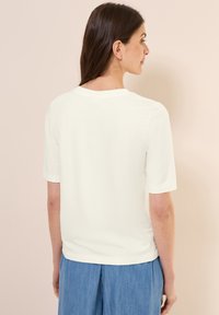 Blouse blanche à manches courtes avec un col rond et une texture lisse. Le mannequin porte un pantalon léger bleu clair, ample.
