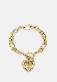 Bracelet en métal doré avec des maillons surdimensionnés, présentant un charm en forme de cœur orné de strass et de détails en logo embossés.