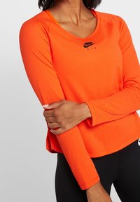 Nike longsleeve top in feloranje met een gestructureerde stof, ronde hals en zwart logo op de borst, gecombineerd met zwarte leggings.