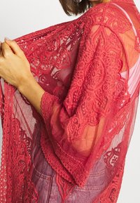 Syväpunaista pitsikimonoa koristavat monimutkaiset kukkakuviot, läpikuultava kangas ja leveät hihat. Reunat ovat aaltoilevat lisäyksinä.