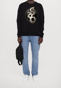 Man som bär en svart sweatshirt med en guldorm och texten "Armani Exchange", ljusblå jeans, vita sneakers, håller en svart necessär.