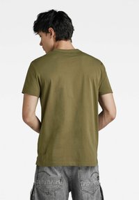 T-shirt in cotone verde oliva con scollo a girocollo e maniche corte, indossata al contrario, abbinata a pantaloni corti grigi casual con tasche e cuciture.