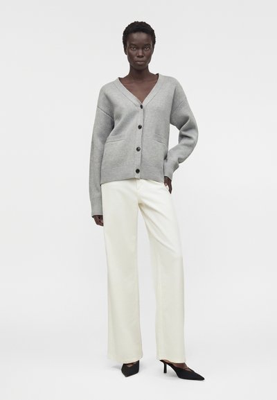 Γυναίκα που φοράει γκρι ζακέτα με κουμπιά, άσπρο παντελόνι φαρδιάς γραμμής και μαύρες μυτερές γόβες slingback, στέκεται μπροστά σε απλό φόντο.