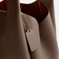 Brown textured leather tote bag with a matching leather tag embossed with brand name, attached by a small gold bead chain.