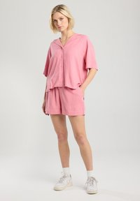 Camisa de lino rosa de manga corta con cuello y botones en el frente, combinada con pantalones cortos de lino a juego; el modelo lleva zapatillas blancas y calcetines.