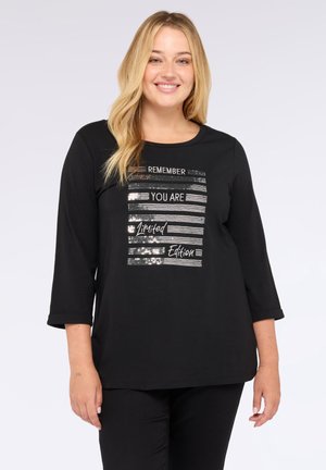 Mujer rubia sonriente con una camiseta de manga larga negra con el texto plateado brillante "Recuerda que eres una edición limitada" en el frente, de pie frente a un fondo blanco.