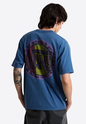 Man met tatoeage op zijn arm, draagt een blauw Vans T-shirt, met de rug naar de kijker, toont een groot kleurrijk Vans-logo en tekst op zijn rug.
