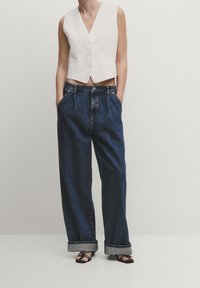 Biała kamizelka bez rękawów z głębokim dekoltem w serek i guzikami, zestawiona z luźnymi, ciemnoniebieskimi dżinsami z mankietami i kieszeniami po bokach.