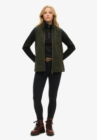 Groene fleece vest met blauwe accenten, gecombineerd met een zwart langemouwen shirt en donkere jeans. Bruine veterschoenen maken de outfit compleet.