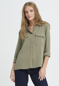 Chemise vert olive à boutons avec deux poches poitrine, manches longues retroussées, texture douce et ourlet arrondi.