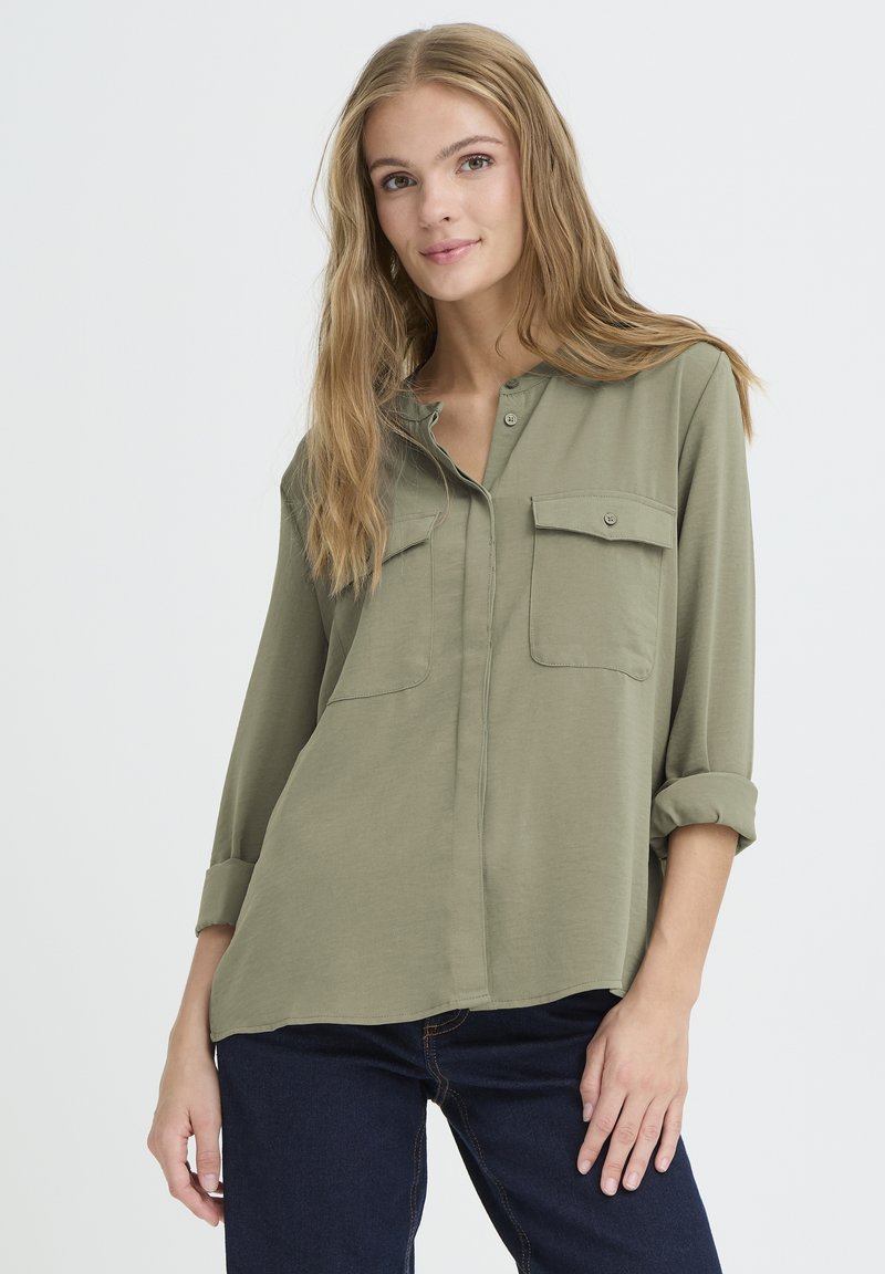 Chemise vert olive à boutons avec deux poches poitrine, manches longues retroussées, texture douce et ourlet arrondi.