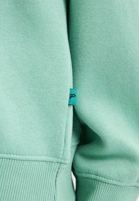 Il tessuto di una felpa verde menta mostra una morbida texture, con polsini a coste e un piccolo etichetta turchese sul lato.