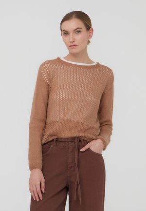 Donna che indossa un maglione marrone chiaro a maglia con maniche lunghe e pantaloni a vita alta marrone scuro con cintura annodata, posando con una mano in tasca.