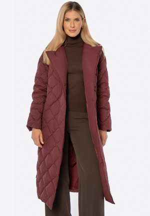 Vrouw met lang blond haar die een bordeauxrode doorgestikte lange jas draagt over een bruine coltrui en bruine wijde broek.