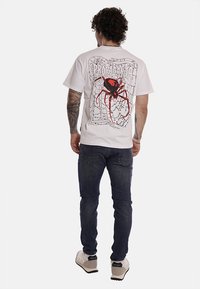 T-shirt bianca in cotone con una grande grafica di un ragno nera e rossa sul retro, scollo tondo e maniche corte, abbinata a jeans scuri.