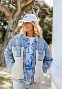 Denimjakke med cremede sherpa-paneler, to brystlommer, sølv hardware, båret over en hvid t-shirt med blåt blomstermønster, lyse baggrund.