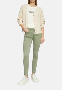 Beige gebreide cardigan over een wit en licht gestreept shirt, gecombineerd met olijfgroen skinny broek en witte platformsneakers.