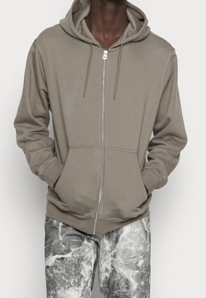 Man wearing taupe zip-up hoodie with drawstrings and front pockets, paired with black and white marble-patterned pants.