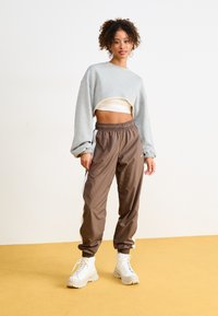Sweatshirt cinza, cortado, com mangas longas, combinado com calças de treino castanhas com riscas brancas nas laterais e ténis brancos robustos.
