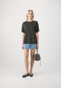 Camiseta corta de manga corta en verde oscuro con tachuelas metálicas dispersas, combinada con una mini falda de mezclilla azul claro y unos zapatos planos negros con tachuelas.