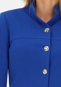 Niebieska kurtka wykonana z gładkiego materiału, z wysokim kołnierzem i pięcioma błyszczącymi srebrnymi guzikami w kształcie promieni słonecznych. Prosty, dopasowany krój.