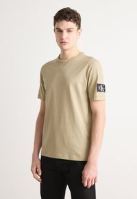Khaki bavlnené tričko s krátkymi rukávmi, okrúhlym výstrihom a čiernou nášivkou na ľavom rukáve. Hladká textúra a bežný strih.