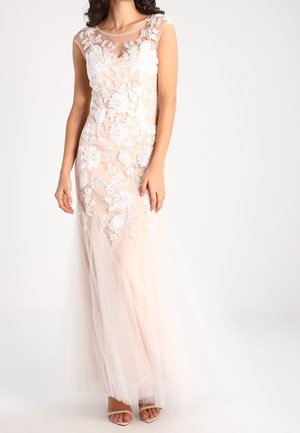 Kobieta w bezrękawnikowej sukni w kolorze różu z białymi kwiatowymi aplikacjami z koronki i przezroczystą warstwą tiulu, połączona z cielistymi szpilkami.