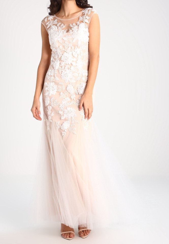 Femme portant une robe rose blush sans manches avec des applications en dentelle florale blanche et une superposition en tulle transparent, associée à des talons couleur nude.