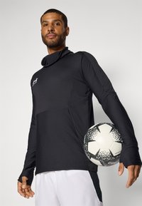 Long-sleeve black jersey with textured fabric, high neck, and thumbholes, paired with a black-and-white soccer ball featuring a speckled pattern.
