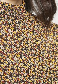 Fließende Bluse mit einem gerafften Ausschnitt. Aus dunkelbraunem Stoff, verziert mit kleinen gelben, roten und weißen Blumenmustern.