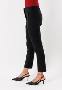 Donna che indossa pantaloni neri su misura e tacchi a slingback neri, in piedi contro uno sfondo bianco, mostrare la vista laterale dalla vita in giù.