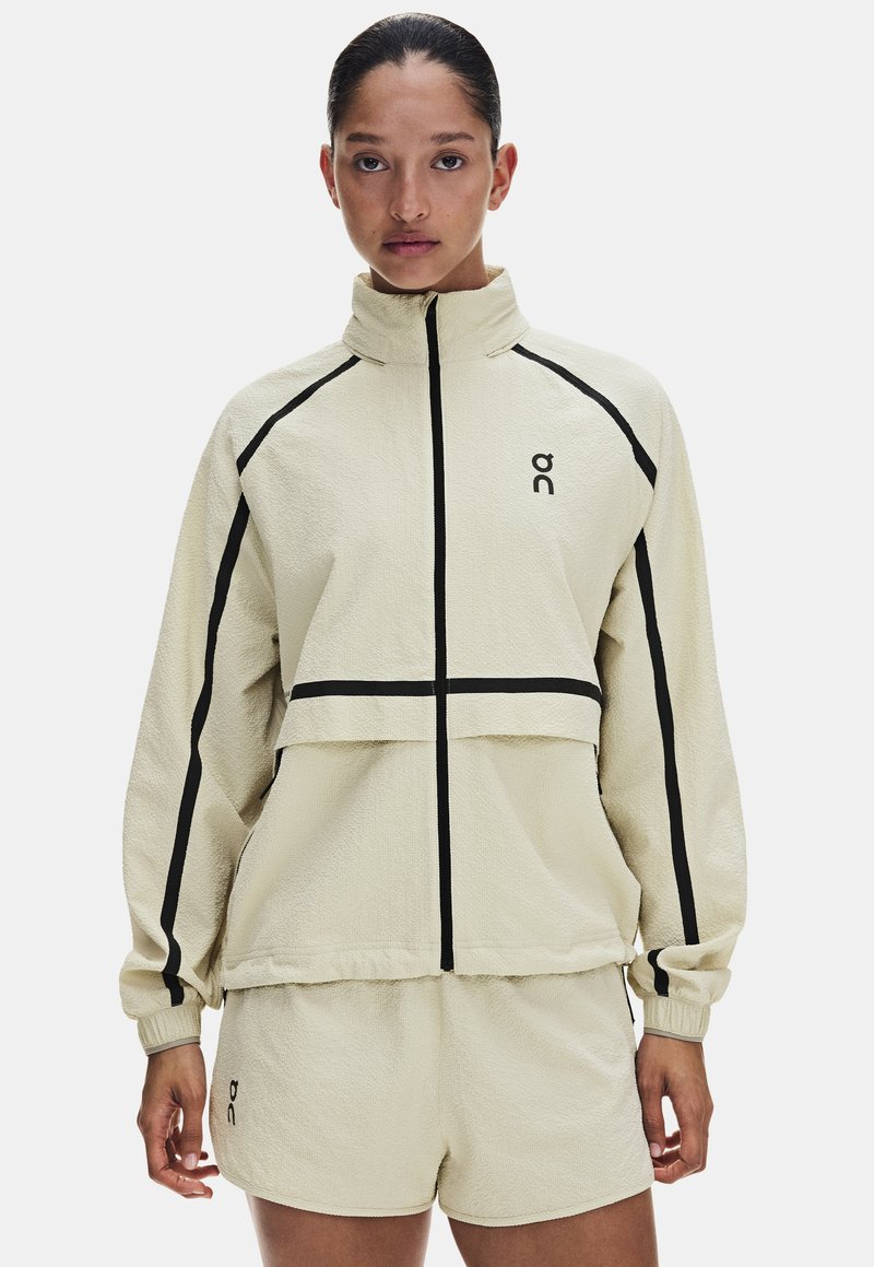 Femme portant une veste beige à fermeture éclair et un short assorti avec des détails en lignes noires et un petit logo sur la poitrine et le short.