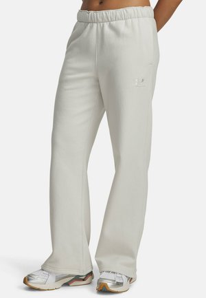 Persoon draagt losse, lichtgrijze joggingbroek met elastische tailleband en zilveren sneakers, staand tegen een effen achtergrond.
