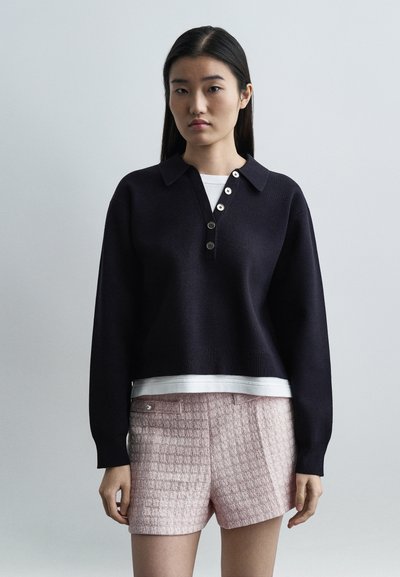 Femme aux cheveux noirs lisses portant un pull noir boutonné par-dessus une chemise blanche et un short rose texturé, debout devant un fond uni.