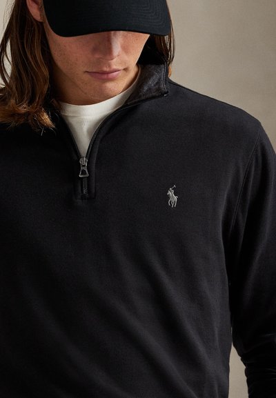 Junge Person trägt schwarzen Viertel-Zip-Pullover mit besticktem Logo auf der Brust und schwarze Kappe, schaut nach unten.