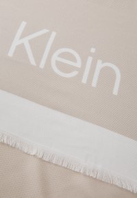 Beige vevd stoff med en teksturert mønster, med en hvit stripe med "Klein" trykt med store bokstaver og frynser på kanten.