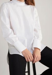 Blusa bianca a maniche lunghe con collare arricciato, texture liscia e maniche gathered. Indossata con pantaloni neri, mani appoggiate su una sedia.