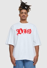 Balta per didelį medvilninį T-shirt su ryškiai raudonais „DIO“ grafikos užrašais. Trumpomis rankovėmis, apvaliu kaklu, švelni tekstūra. Dėvima su mėlynais džinsais.