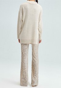 Světlobeige oversize svetr spárovaný s třpytivými flared kalhotami, které mají jemnou texturu a boční rozparky na lemu.