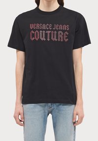 Černé bavlněné tričko s výrazným nápisem "Versace Jeans Couture" v červené a stříbrné barvě v moderním písmu na hrudi. Krátké rukávy.