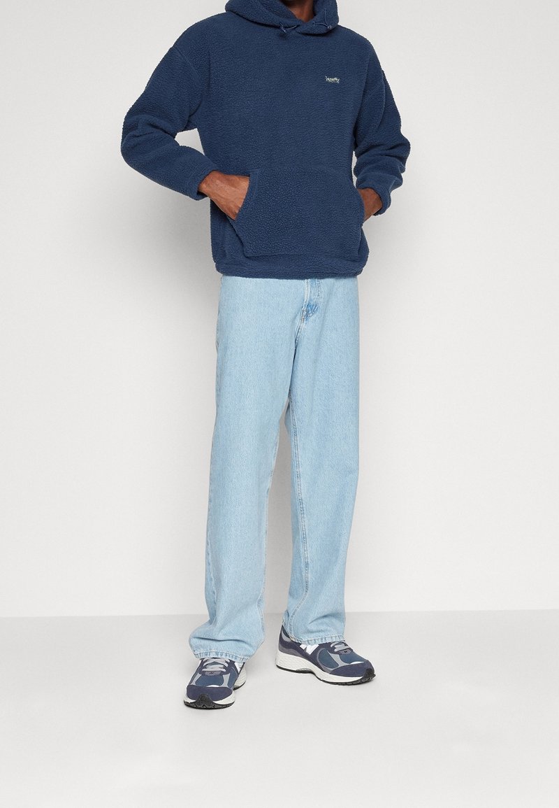 Felpa con cappuccio in pile blu navy con tasca a marsupio, abbinata a jeans azzurri e sneaker blu navy con dettagli grigi e un design testurizzato.