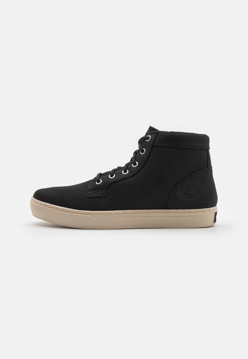 Basket noire montante avec semelle en caoutchouc beige, lacets sur le devant et logo embossé sur le côté du talon, présentée sur un fond uni.