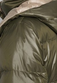 Chaqueta de plumas verde oliva con textura brillante, forro beige en contraste y diseño acolchado. Presenta un cuello alto y puños elásticos.